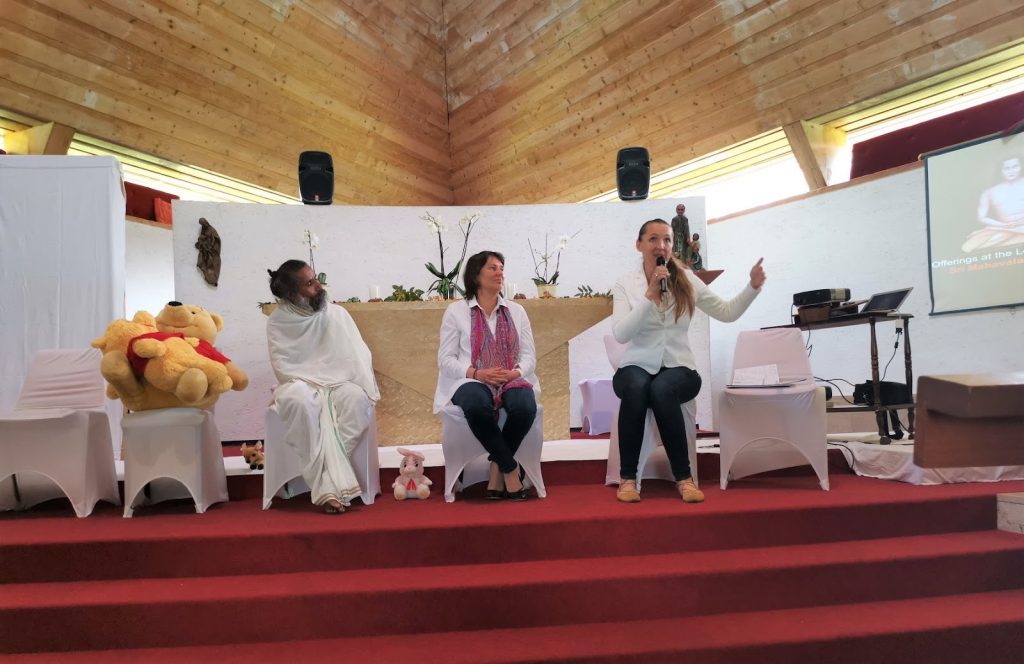 Three women seated on a stage, each engaged in conversation, with a teddy bear placed prominently between them.